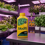 A yellow bottle of Roots Bloom Hydroponic Plant Food positioned on a stainless steel table inside a modern indoor garden facility with purple LED grow lights and potted plants in the background.